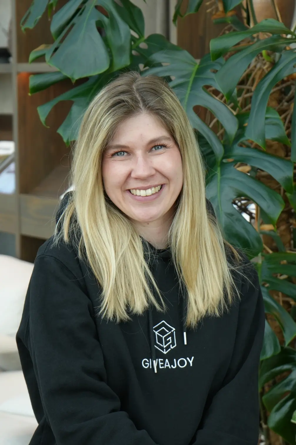 Lächelnde Frau mit langen blonden Haaren trägt schwarzen Hoodie mit GIVEAJOY-Aufschrift.