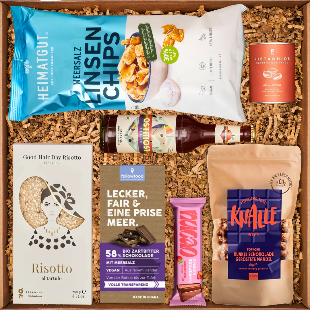 Geschenkbox mit Linsenchips, Risotto, Schokolade, Pistazien, Popcorn und Getränk in braunem Füllmaterial.