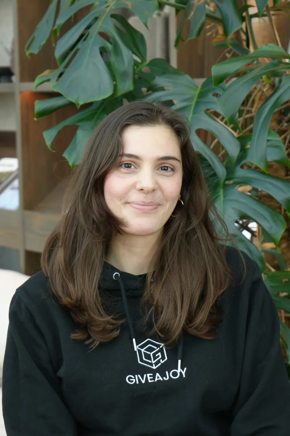 Lächelnde junge Frau mit langen braunen Haaren in einem schwarzen GIVEAJOY Kapuzenpullover vor grünen Pflanzen.