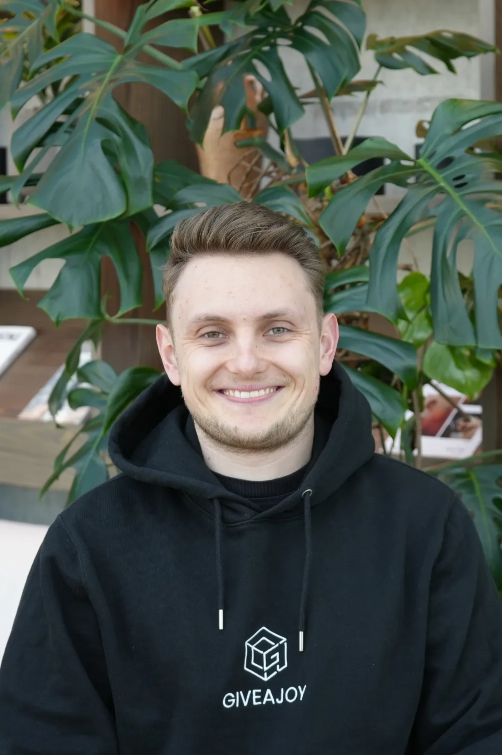 Junger Mann mit blondem Haar lächelt, trägt schwarzen Hoodie mit GIVEAJOY-Logo vor großen grünen Pflanzen.