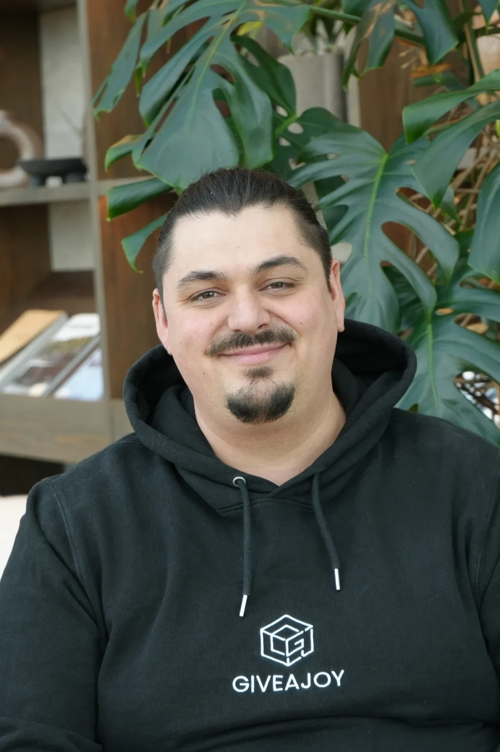 Mann mit Zopf und schwarzem Hoodie vor großen grünen Blättern, lächelnd.