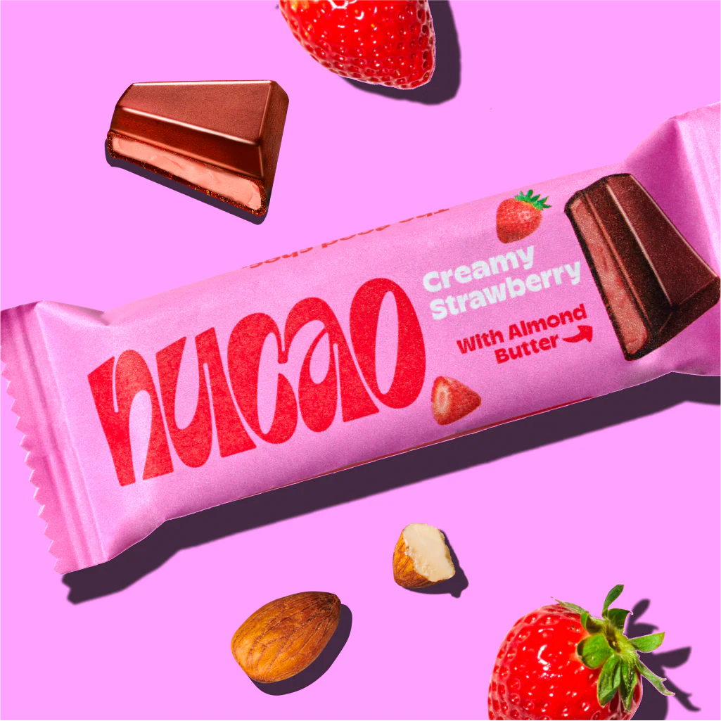 Nucao Creamy Strawberry Riegel mit Mandelbutter, umgeben von Erdbeeren und Mandeln.